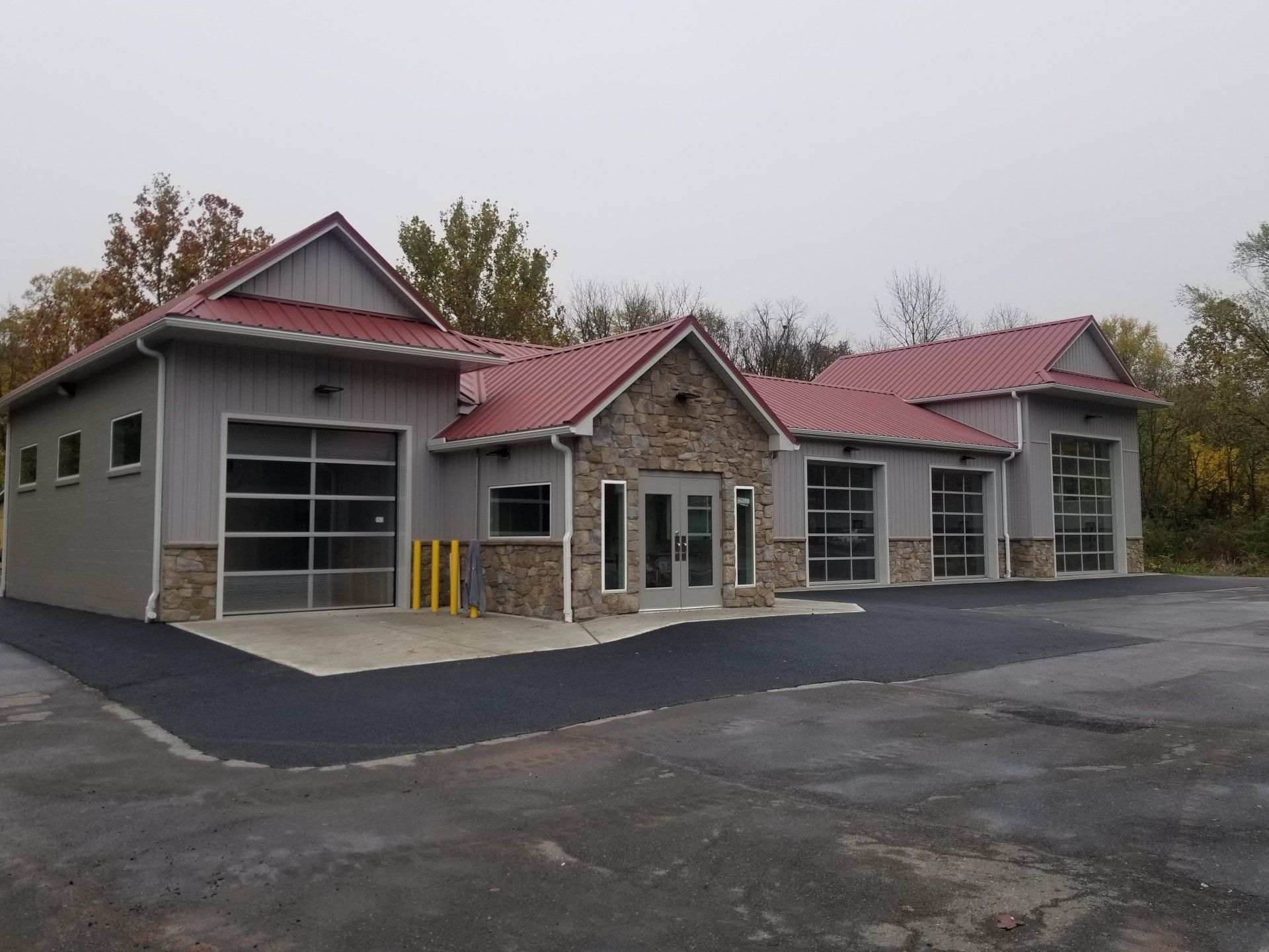 Gray and stone building with red roof, glass garage doors, and a black asphalt driveway on a cloudy day.