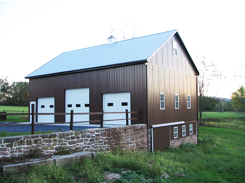 Brown metal barn with white garage doors and a stone foundation on a green landscape.