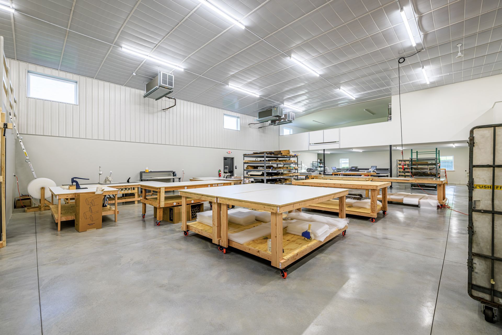 Large workshop with white walls, gray floor, multiple work tables, and storage racks.