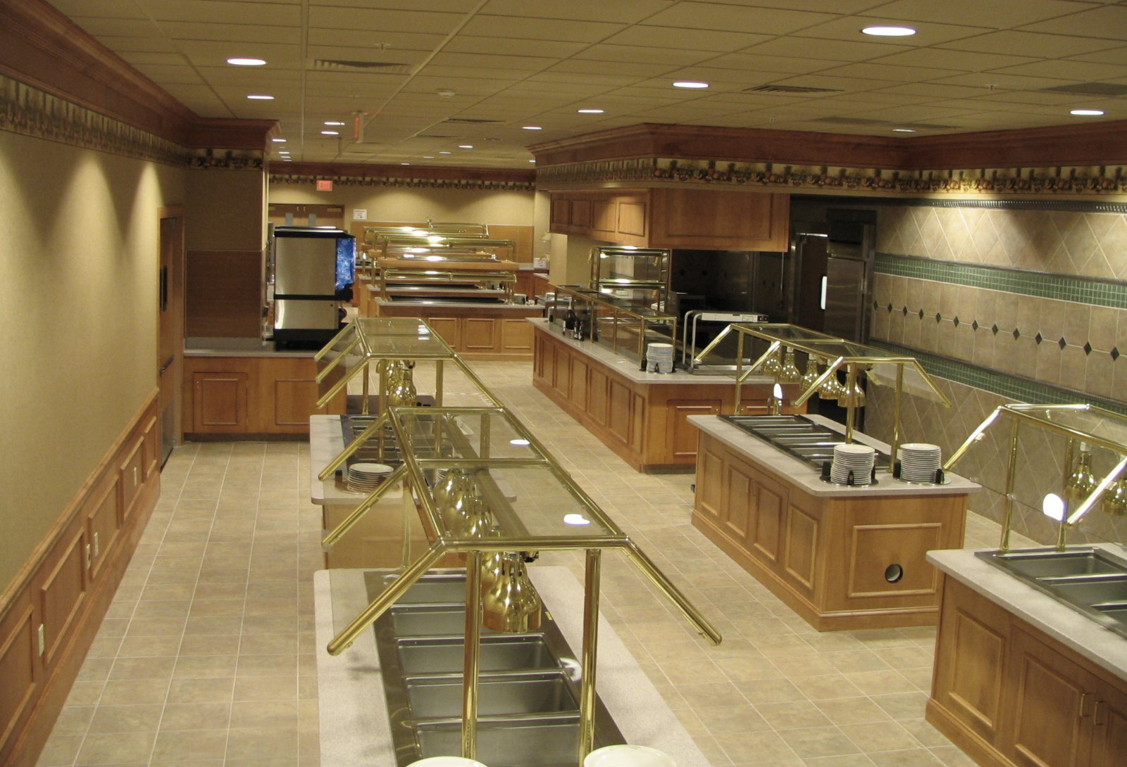 Buffet-style restaurant with food service stations. Light wood cabinets, brass rails, tiled floor, and tan walls.