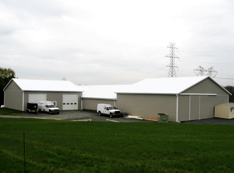 Gray industrial buildings with white roofs and vans parked outside, set in a field under a cloudy sky.
