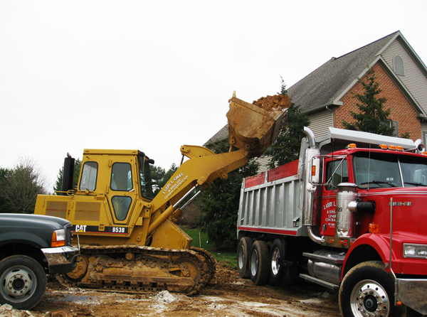 A yellow bulldozer loads dirt into a red dump truck near a house on a cloudy day.