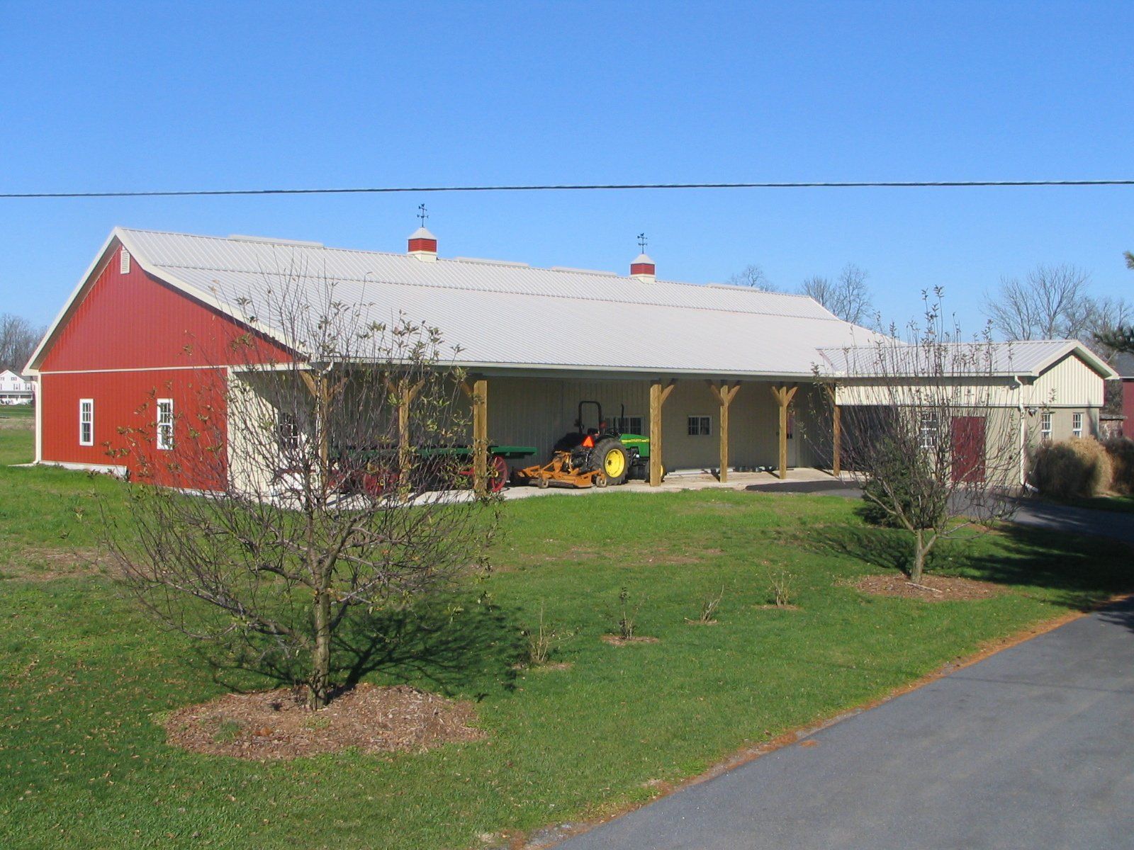 Red and beige barn with metal roof; tractor parked in the covered area, green grass and small trees in front.