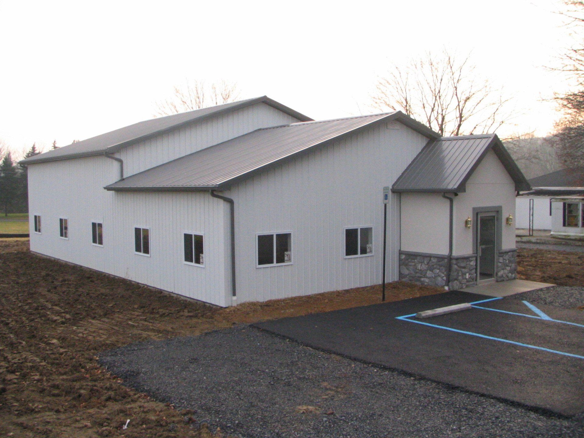 White metal building with a dark roof and a small stone facade entrance; asphalt parking area.
