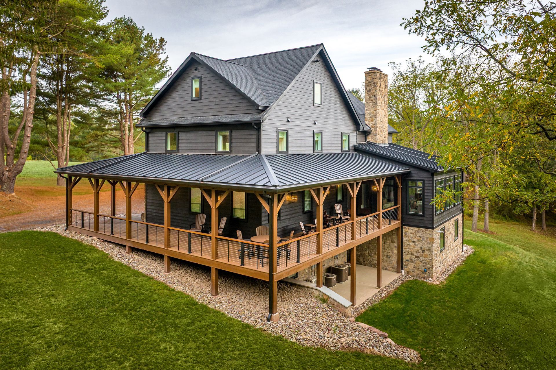 Two-story house with a wraparound porch on a grassy hill. Charcoal gray siding, brown porch, and a stone chimney.