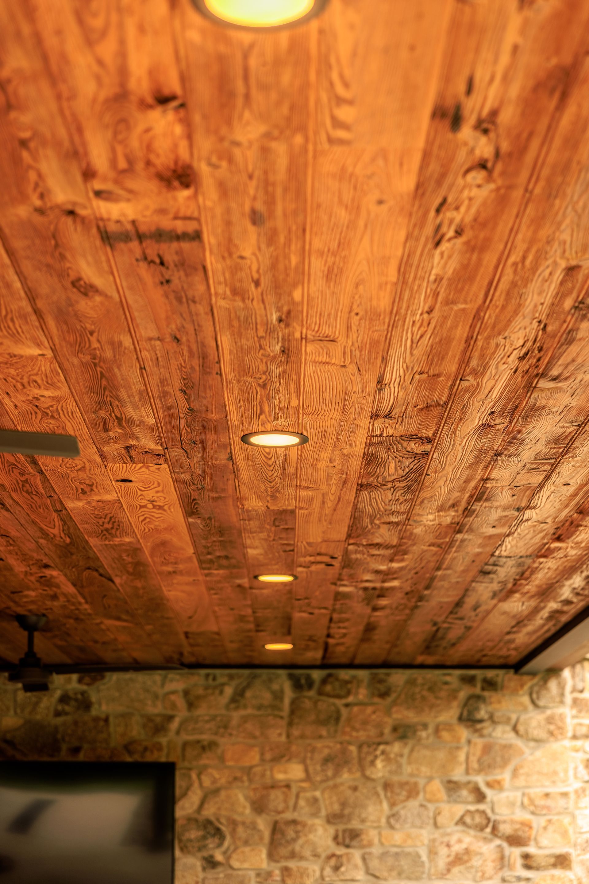 Wooden plank ceiling with recessed lights, above a stone wall. Warm, rustic interior.