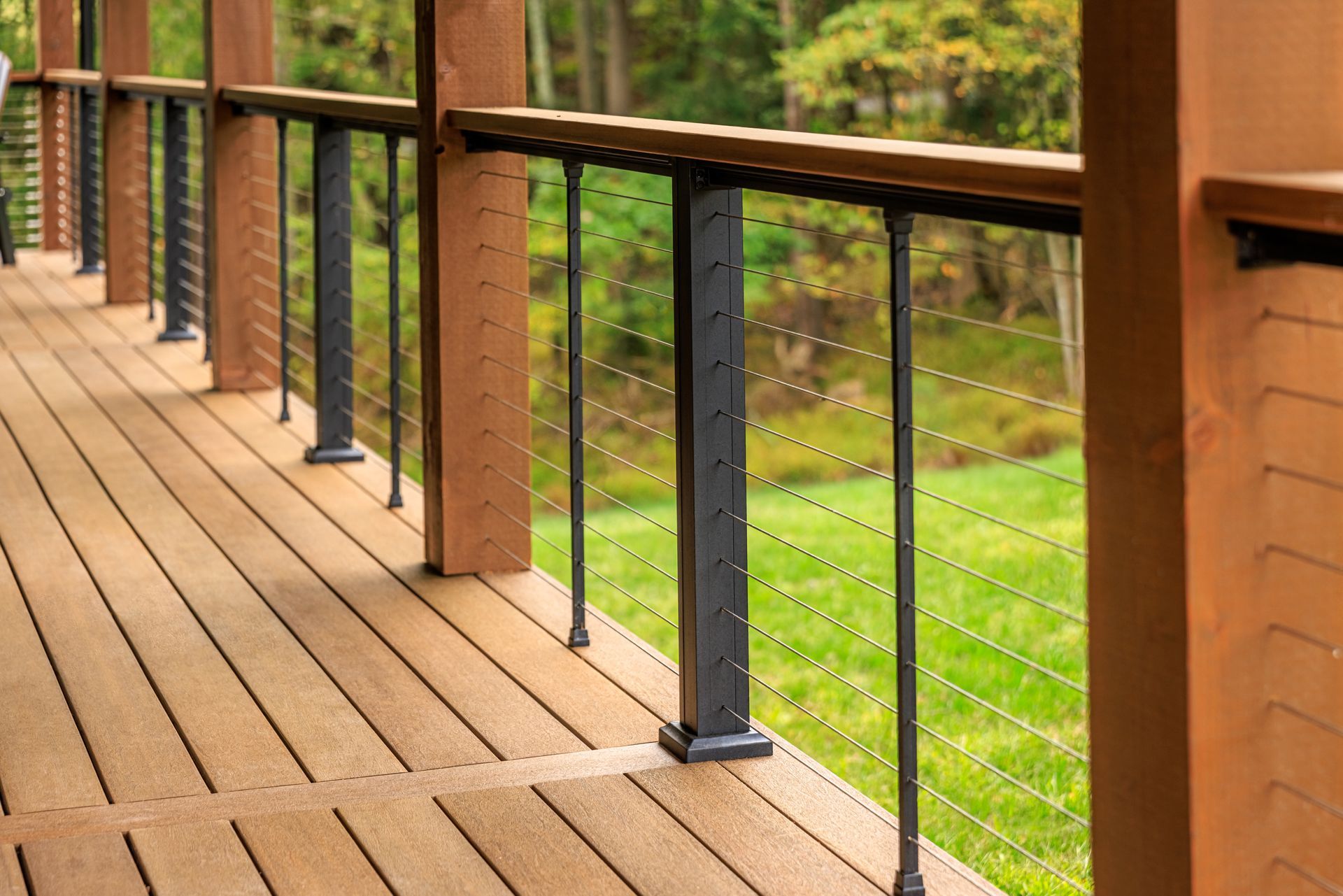 Wooden deck with black metal posts and cable railings overlooking a grassy area.
