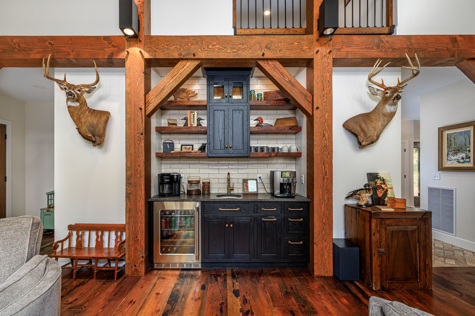 Rustic bar area with black cabinets, open shelving, and deer mounts. Wooden beams and floors.
