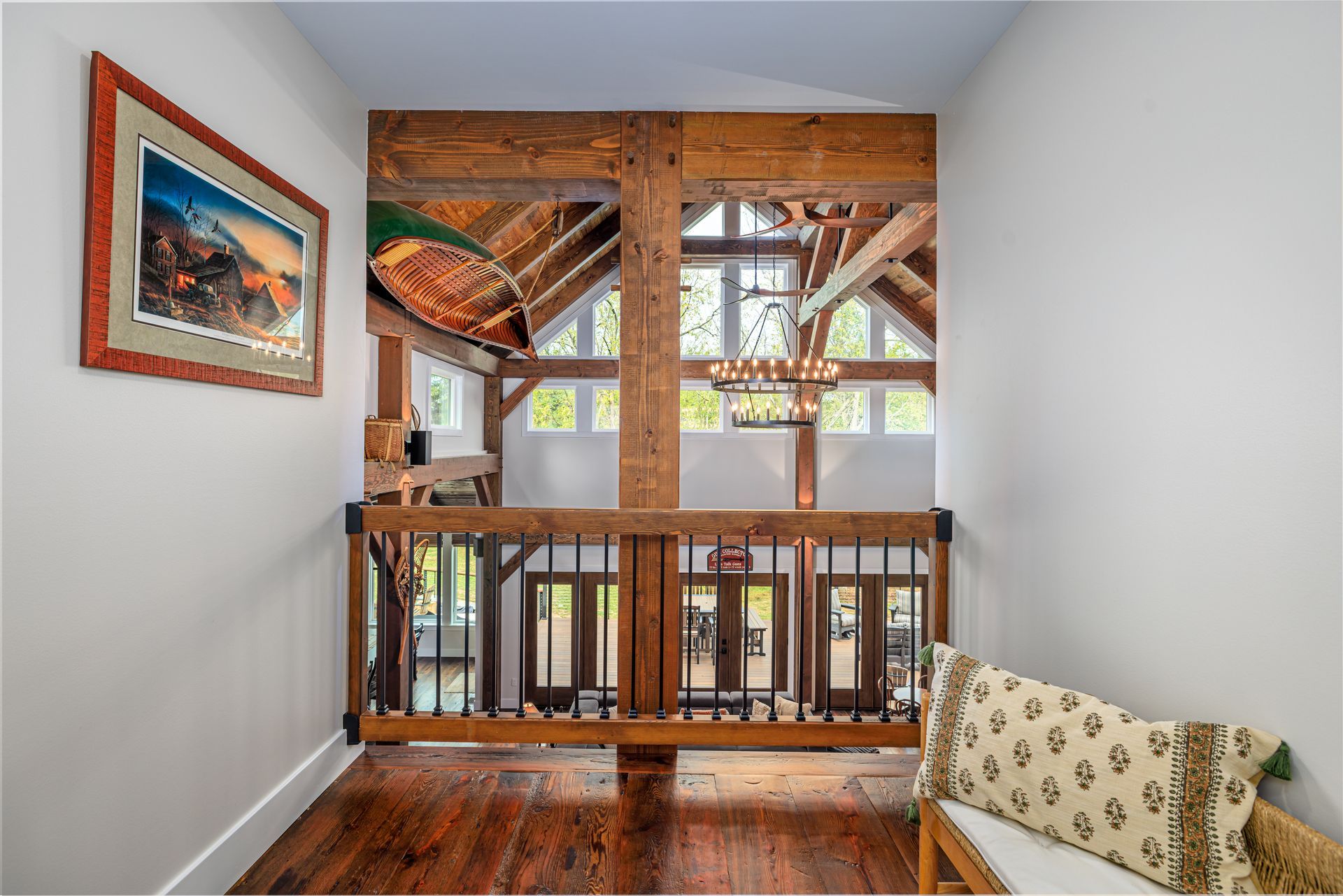 Hallway with rustic wooden beams and railing, overlooking a living space with French doors and a chandelier.