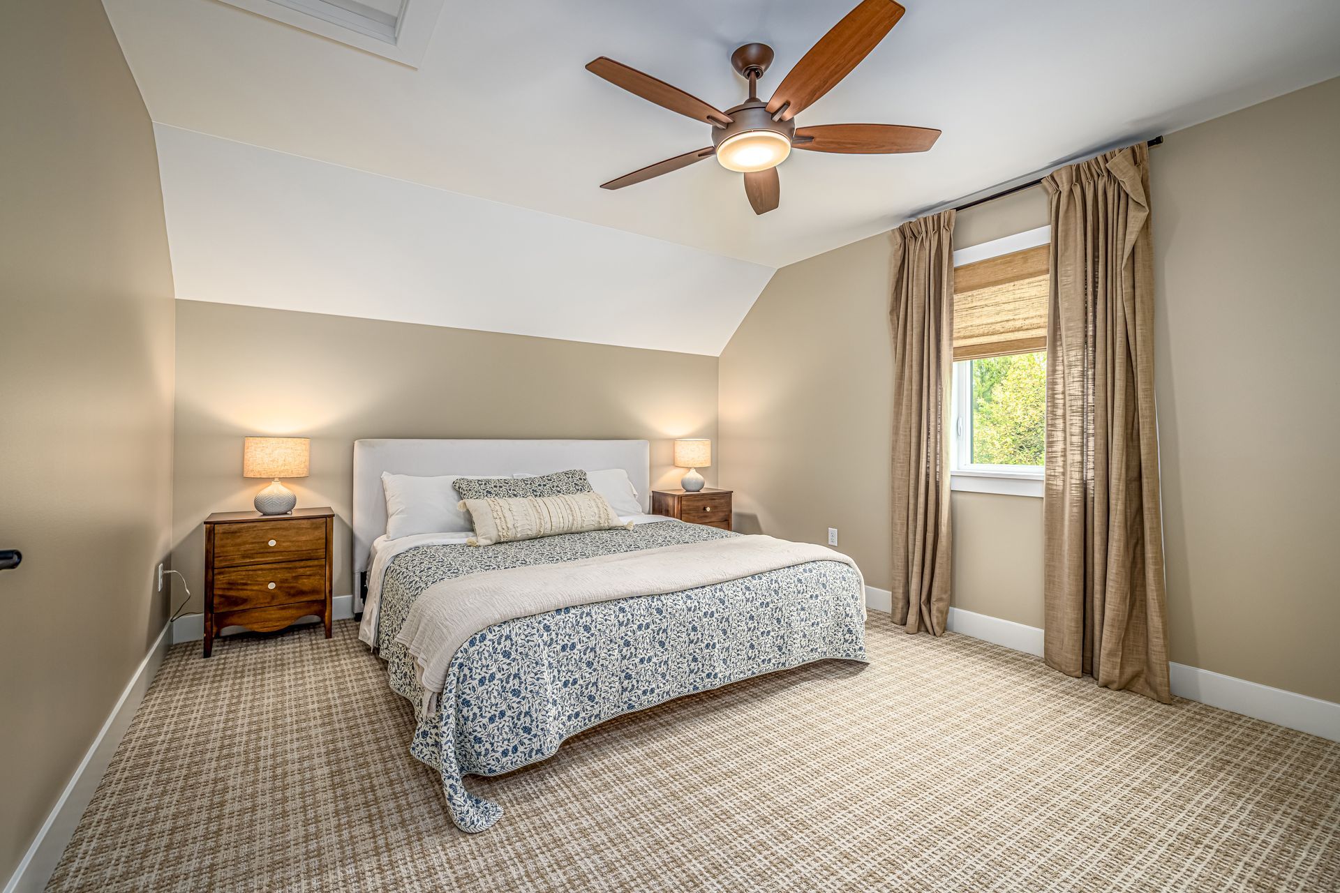 Bedroom with beige walls, cream-colored carpet, bed, nightstands, window with drapes, and a ceiling fan.