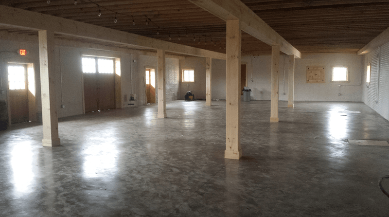 Interior of a large room with concrete floor, wooden beams, and columns; windows and doors on the sides.