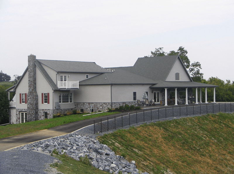 Large, light gray house with stone chimney and front porch, on a hill overlooking a gray fence and grassy area.