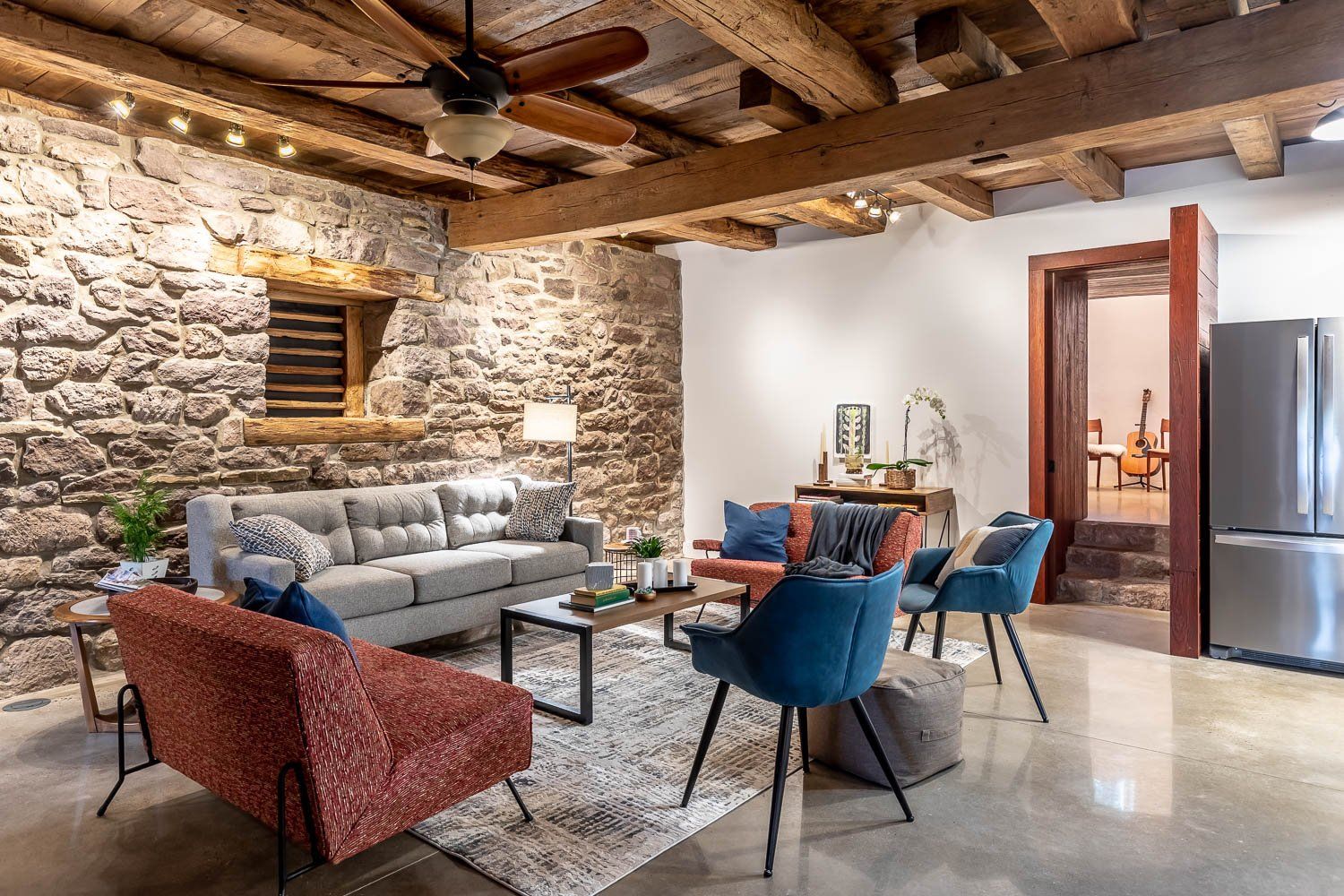 Living room with stone walls, wooden beams, various modern furniture, and a concrete floor.