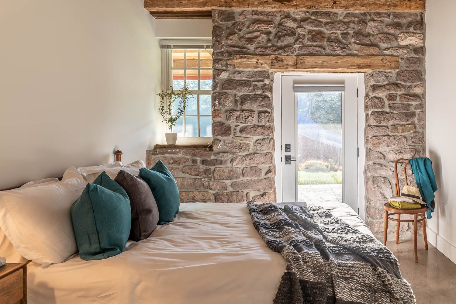Bedroom with stone walls, white bedding, teal pillows, and an open door to a backyard.