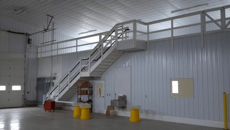 Interior of a white industrial building with a staircase leading to a raised platform, yellow barrels on the floor.
