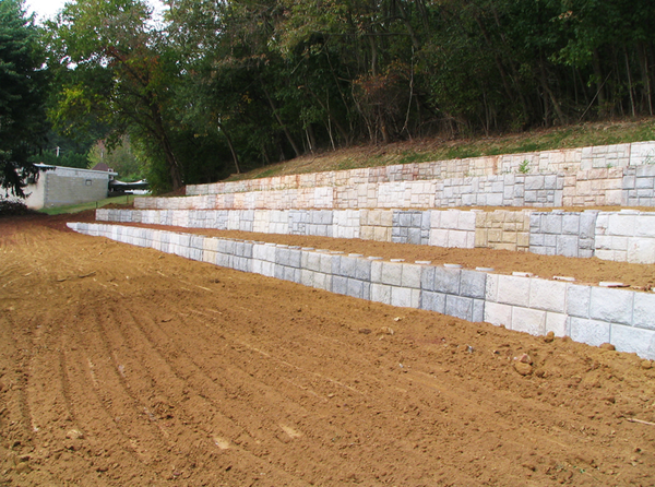 Retaining walls made of concrete blocks on a hillside, with graded dirt in front, trees in the background.