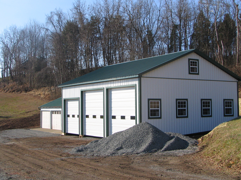 White and green barn with three garage doors and small windows, on a gravel lot near trees.