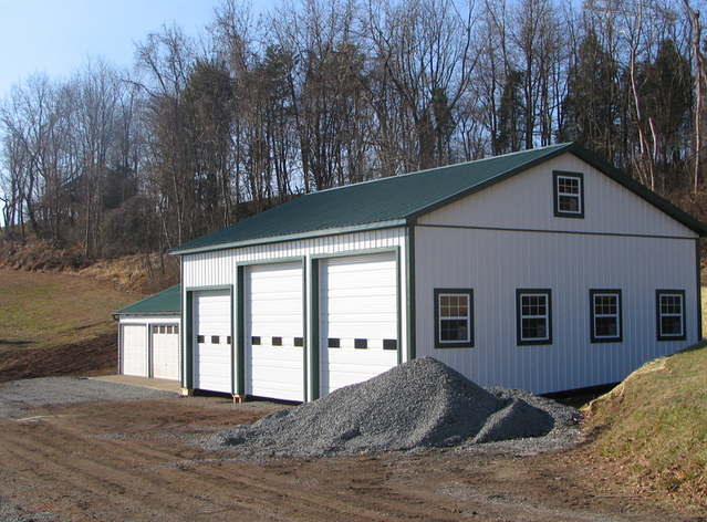 White barn with green trim, three garage doors, windows, and a gravel pile outdoors.
