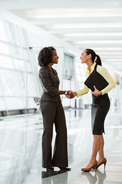 Two women in business attire shaking hands in a bright hallway.