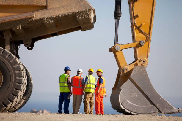 Construction workers in safety vests and hard hats stand near heavy machinery.