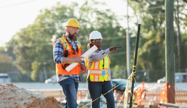 Two construction workers in safety vests and hard hats at a construction site; one points, other holds plans.