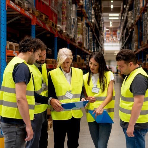 Warehouse workers in safety vests review documents in a busy storage facility.