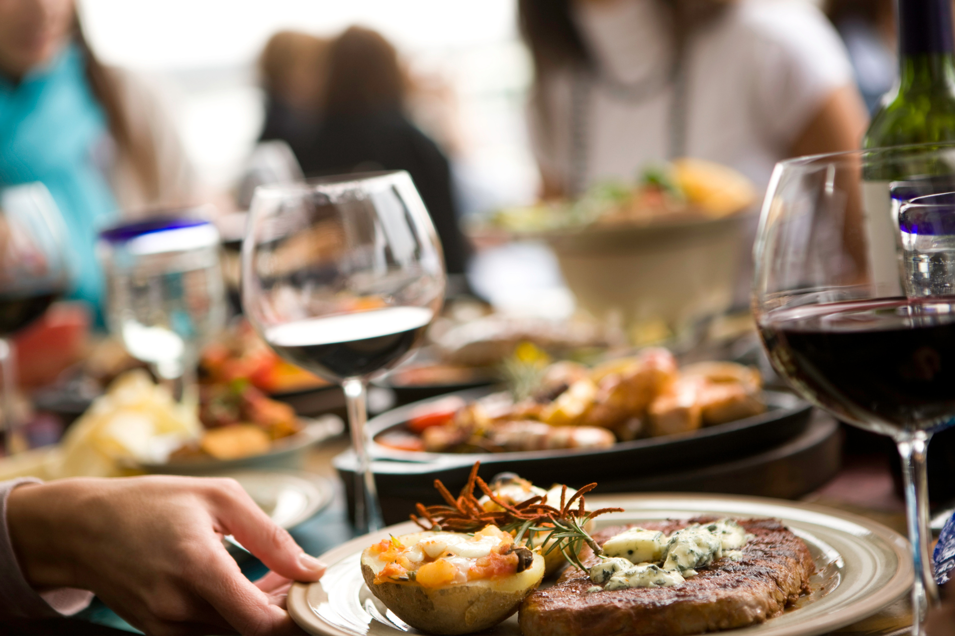 Table with food and drinks, a person holding a plate with a steak and potato. Other people are seated.