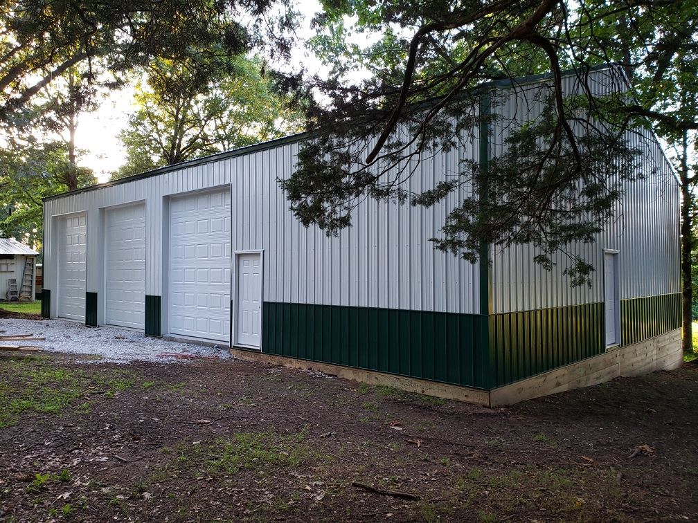 White metal garage building with three roll-up doors, green lower trim, beside trees on a gravel lot. | C & C Automotive