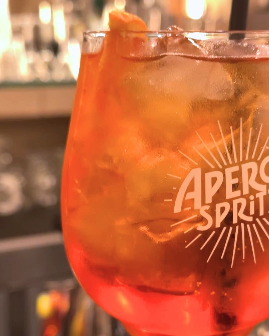 Primo piano del cocktail Aperol Spritz in un bicchiere con ghiaccio e guarnizione all'arancia, illuminato di arancione.