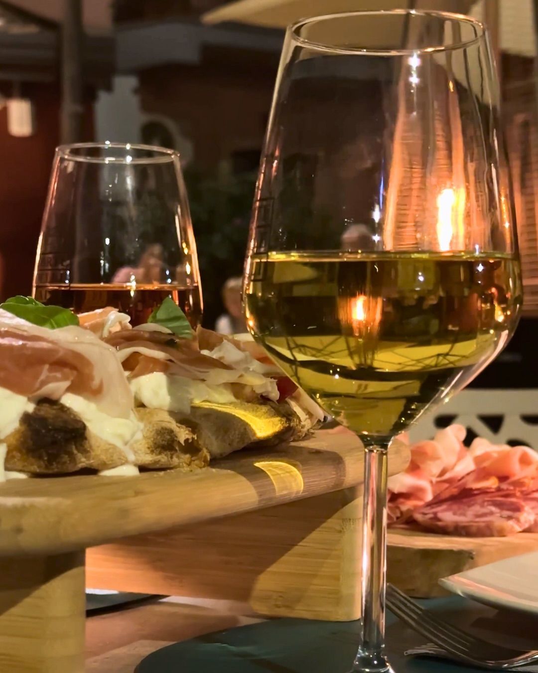 Calici da vino, pizza e salumi su un tagliere di legno. Il caldo bagliore di una fiamma si riflette in un bicchiere.