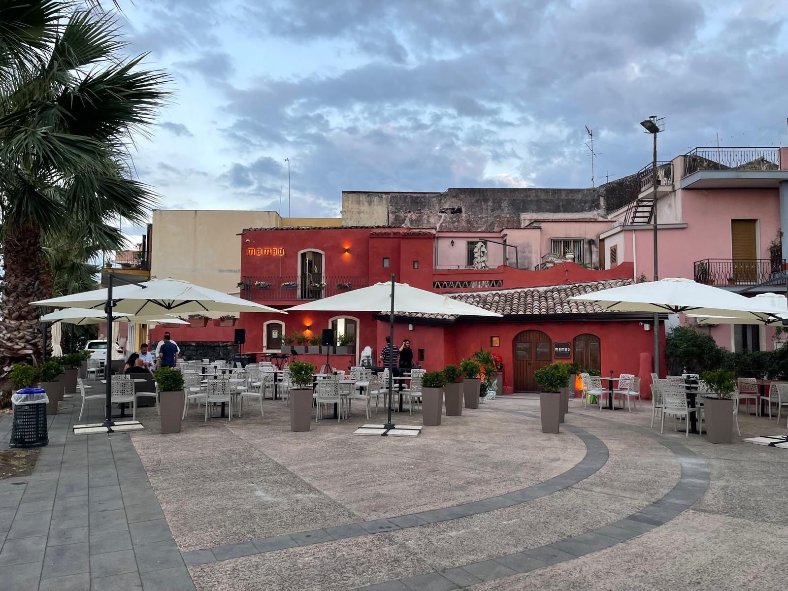 Ristorante all'aperto con edificio rosso, ombrelloni bianchi, tavoli e persone. Piazza acciottolata. Cielo nuvoloso.