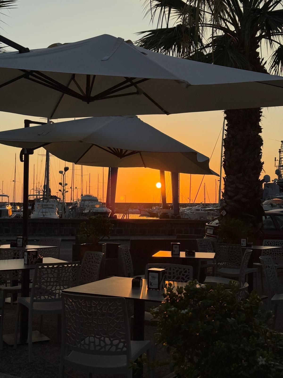 Tramonto sul porto, ammirato dal patio di un ristorante con ombrelloni e tavoli.