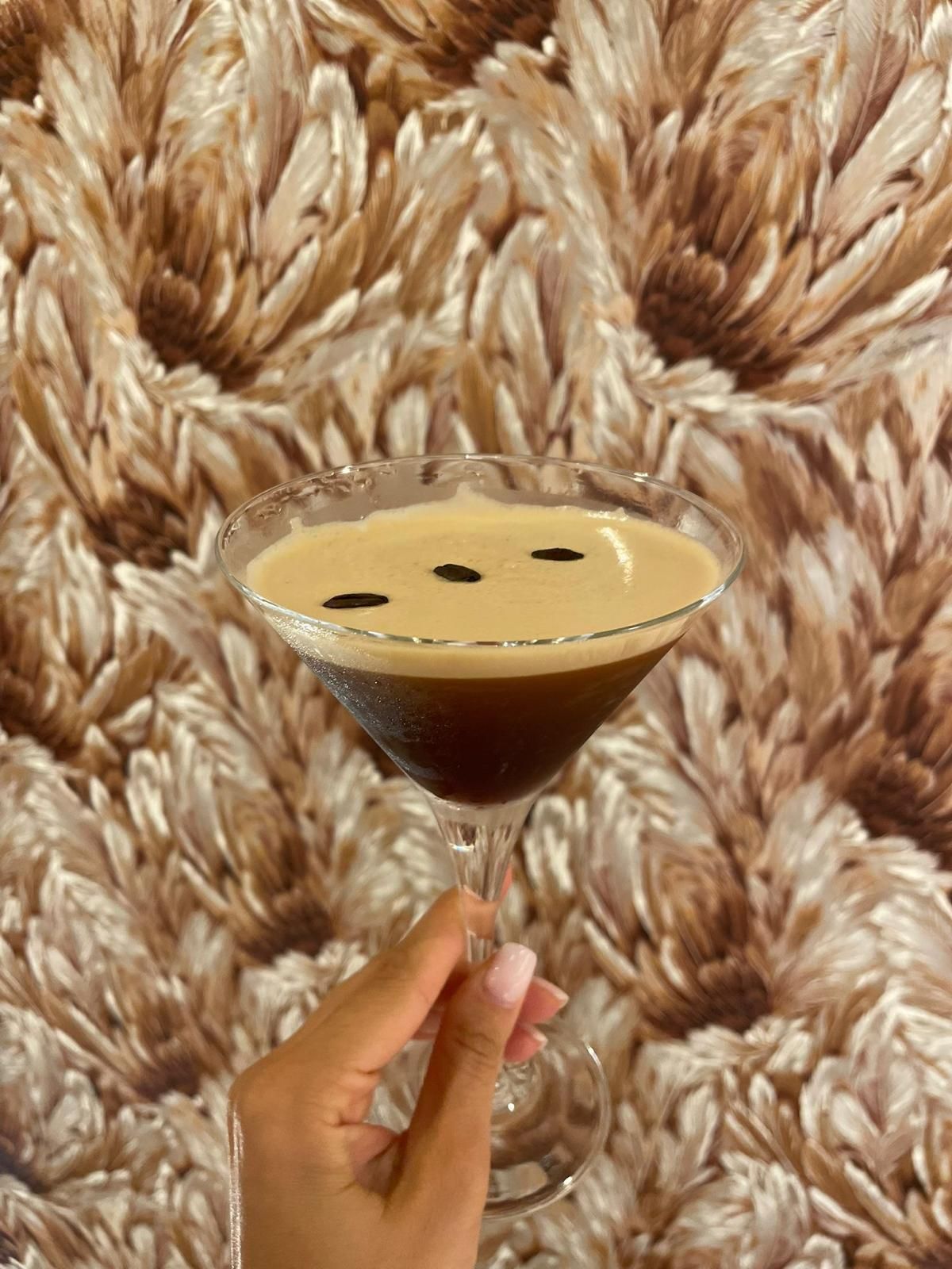 Mano che regge un bicchiere da martini con una bevanda color caffè, su uno sfondo floreale.