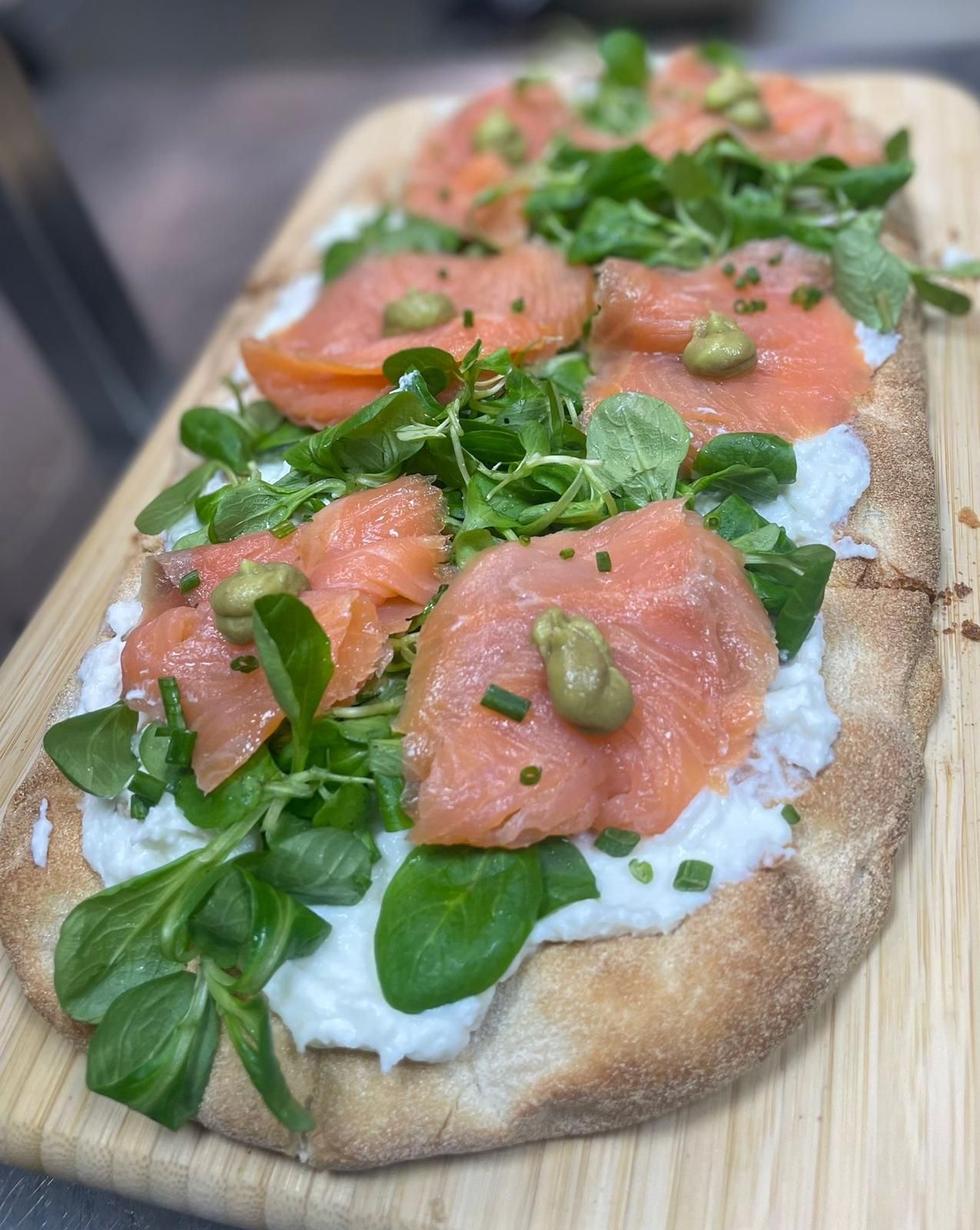 Pane piatto con salmone affumicato, verdure e formaggio cremoso, disposto su un tagliere di legno.