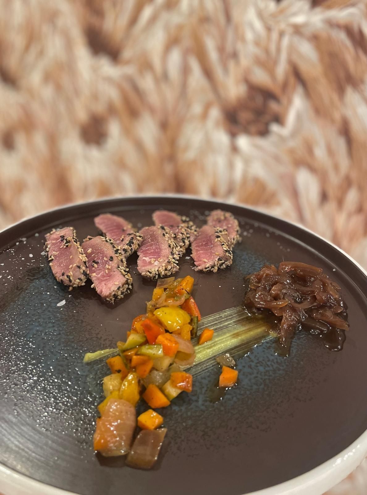 Fette di tonno scottato in crosta di sesamo con verdure saltate e cipolle caramellate su un piatto scuro.