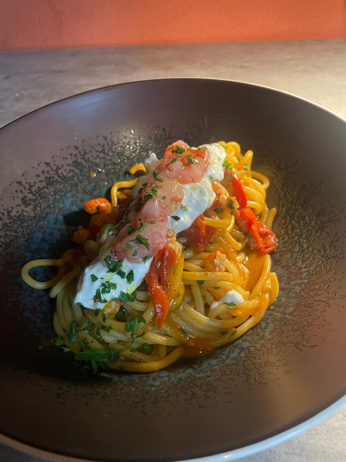 Piatto di spaghetti al sugo di pomodoro, conditi con formaggio cremoso, gamberi ed erbe aromatiche in una ciotola scura.