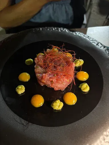 Tartare di tonno su piatto nero, guarnita con puntini di salsa e fili rossi.