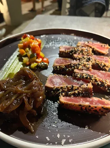 Tonno scottato a fette con semi di sesamo, accompagnato da salsa di verdure e cipolle caramellate, servito su un piatto.