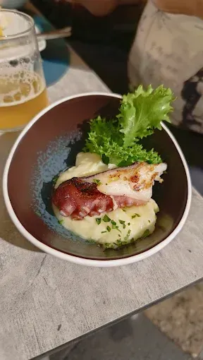 Ciotola di polpo e purè di patate condita con lattuga, su una superficie ruvida, con birra sullo sfondo.