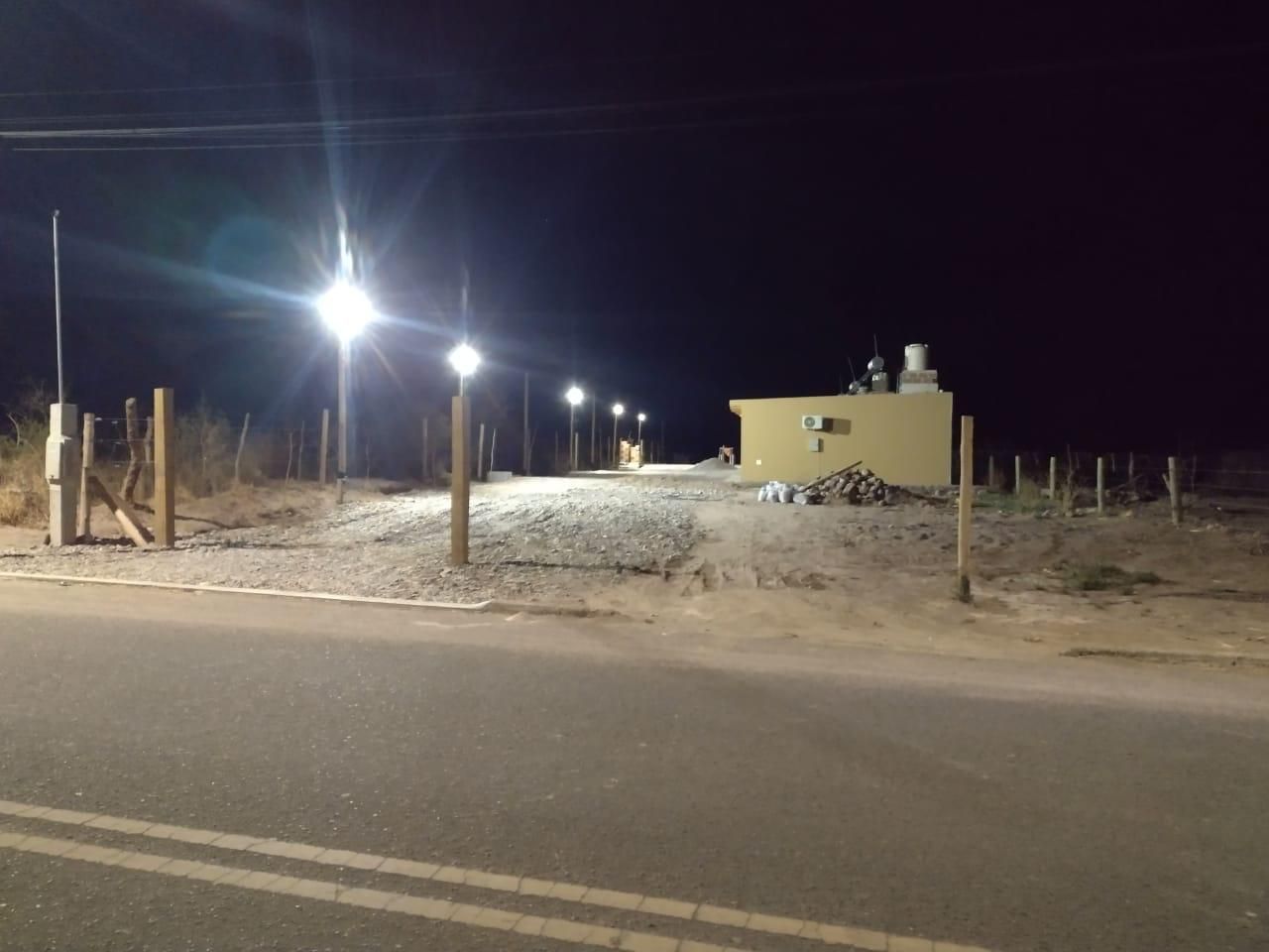 Escena nocturna: camino iluminado que conduce a un edificio beige con estructuras de servicios públicos; postes de madera bordean el camino, cielo oscuro.