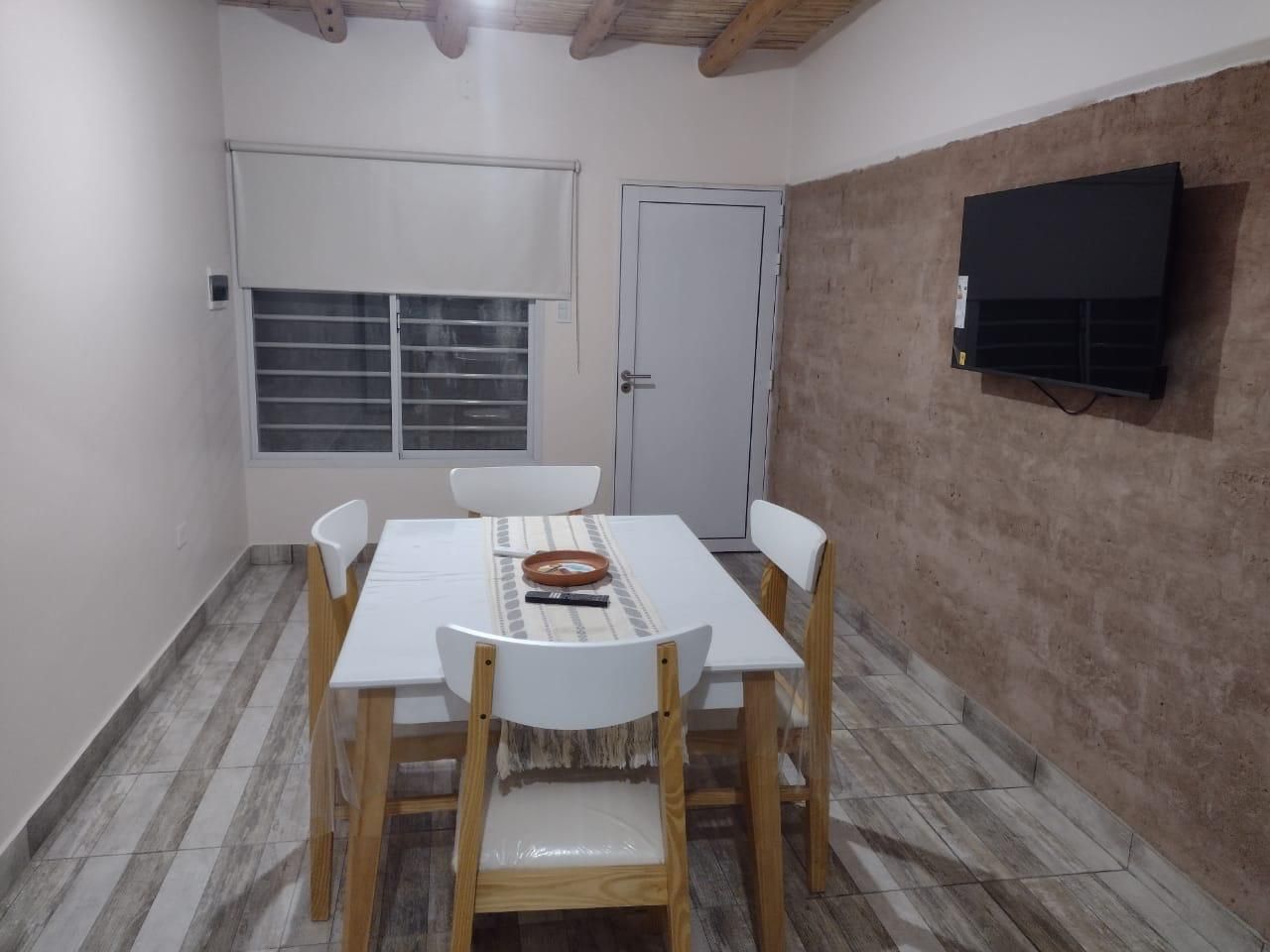 Comedor con mesa blanca, sillas, TV en pared rústica, ventana y puerta.