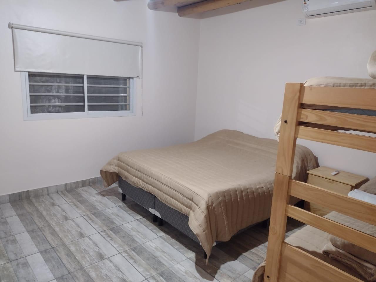 Dormitorio con cama y litera de madera, ropa de cama de color beige y ventana con persiana cerrada.