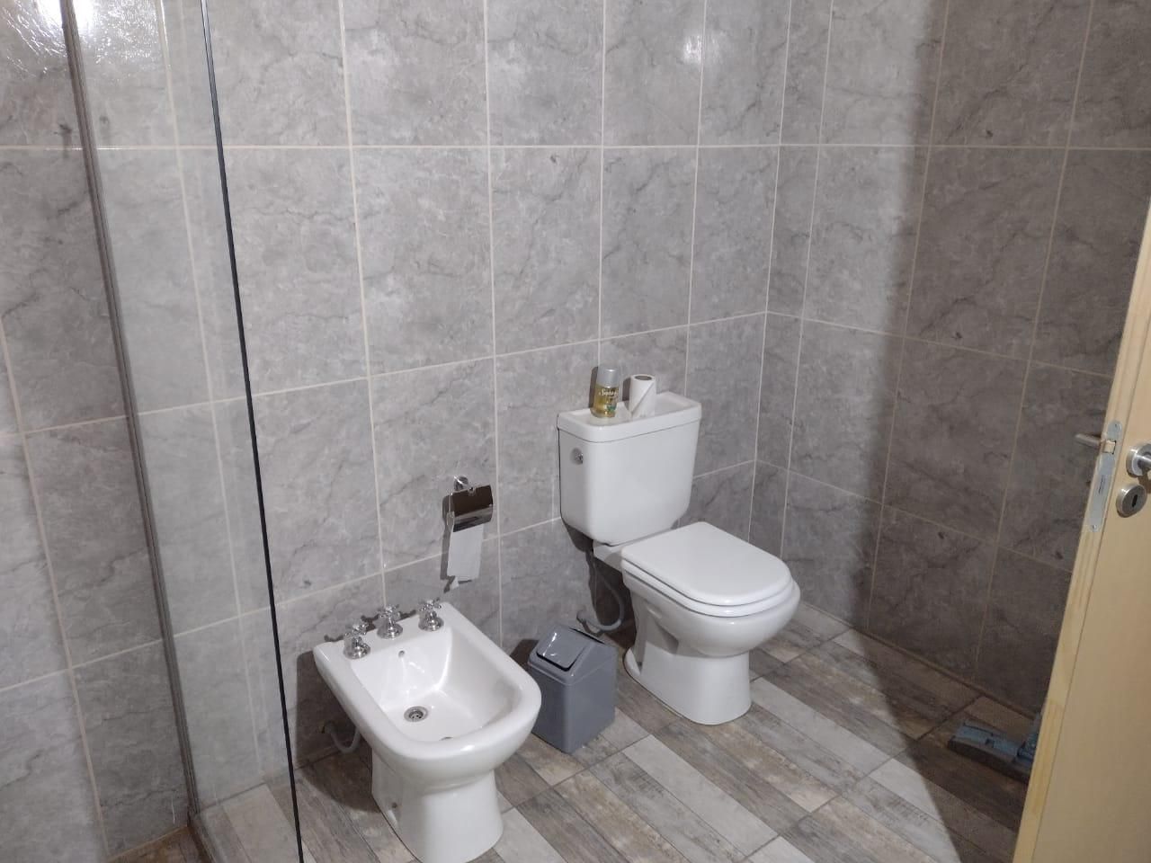 Baño con inodoro, bidé y paredes alicatadas. Tonos grises. Puerta color canela claro a la derecha.