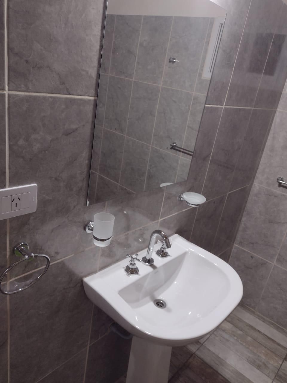 Lavabo de baño con espejo, paredes de azulejos grises y jabonera.