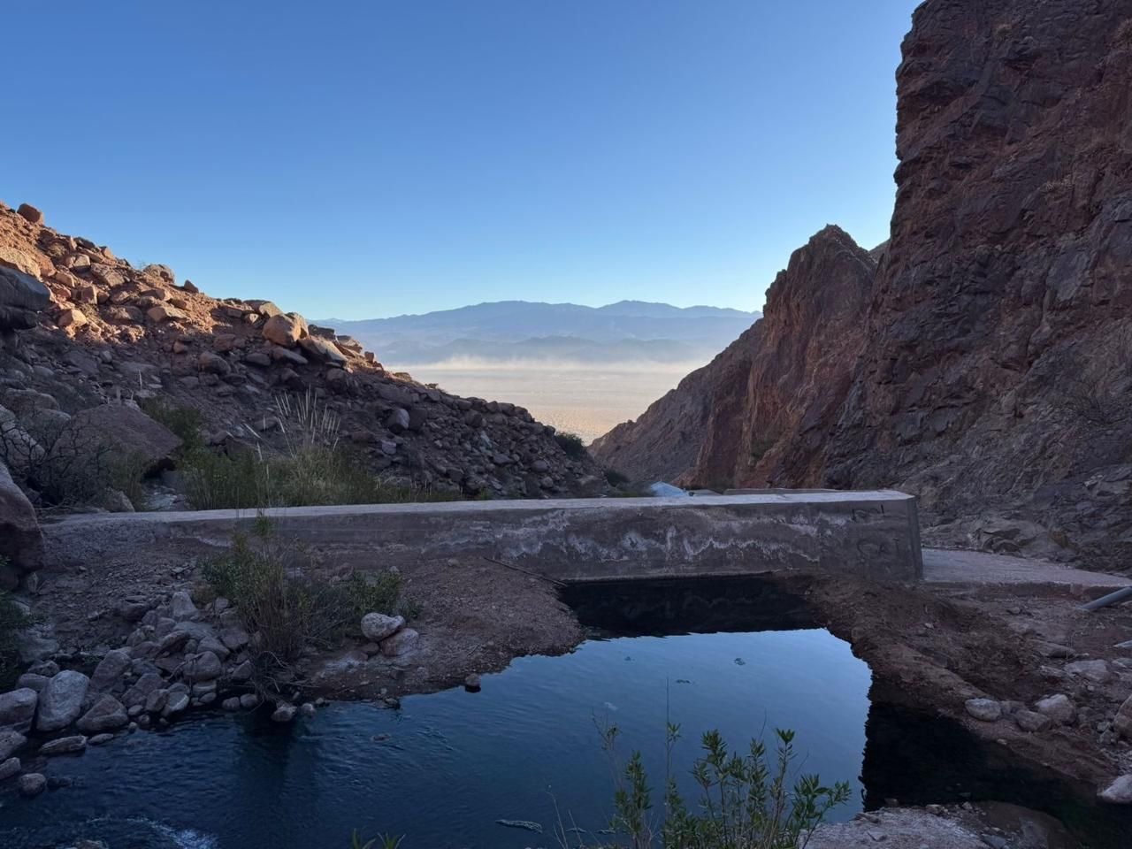 Un puente de hormigón sobre una piscina tranquila en un cañón rocoso, con una cadena montañosa brumosa en la distancia.
