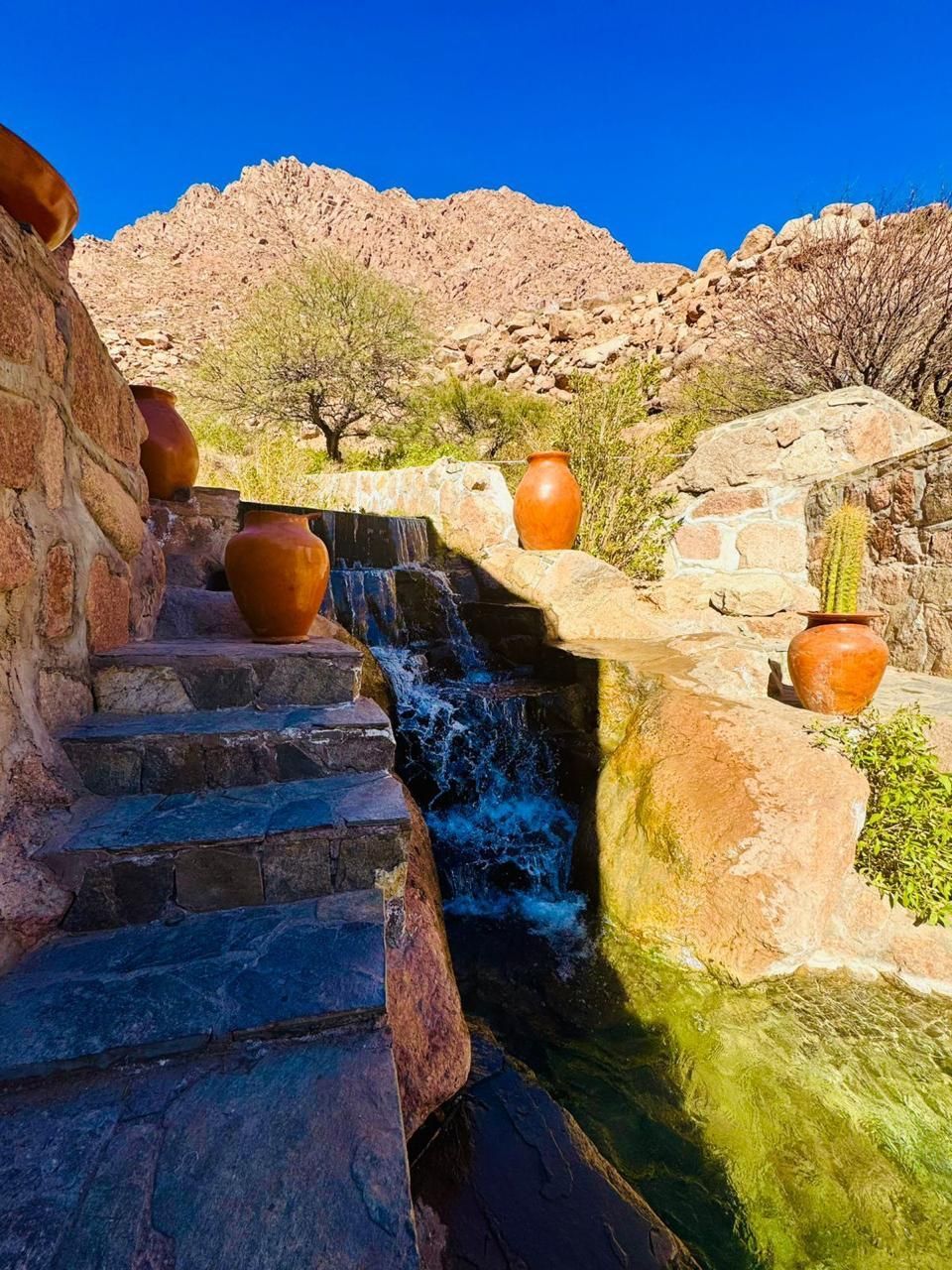 Escalones de piedra y una pequeña cascada fluyen entre vasijas de barro, con un fondo de montañas bajo un cielo azul claro.