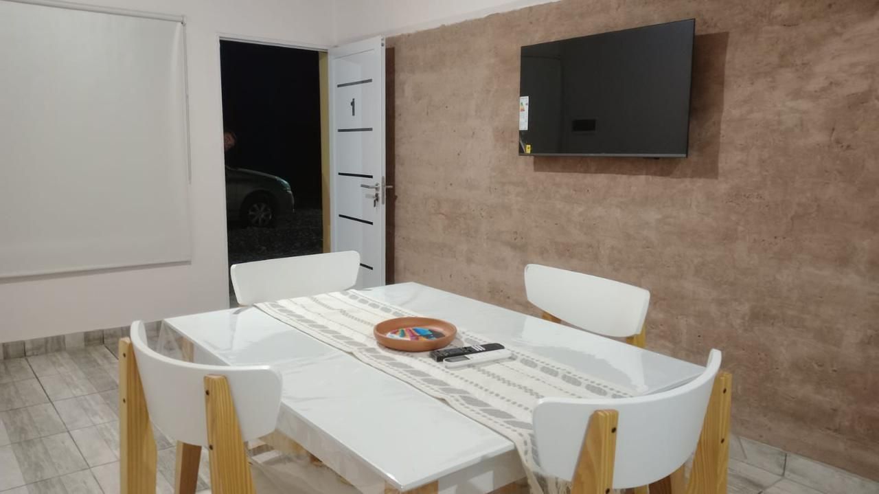 Comedor con mesa y sillas blancas, TV montada y puerta abierta al exterior.