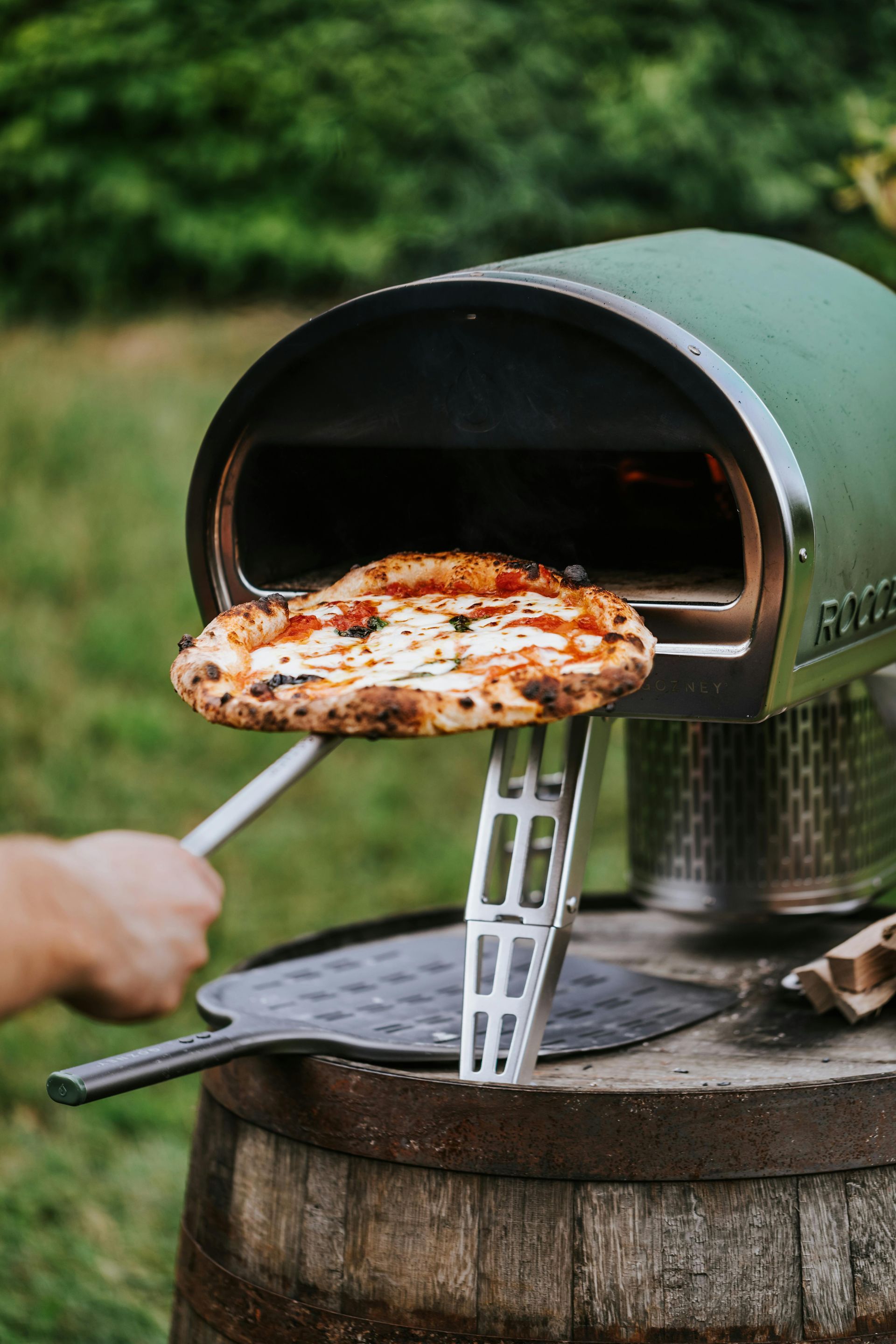 Person removing pizza from a green outdoor pizza oven, set on a barrel.