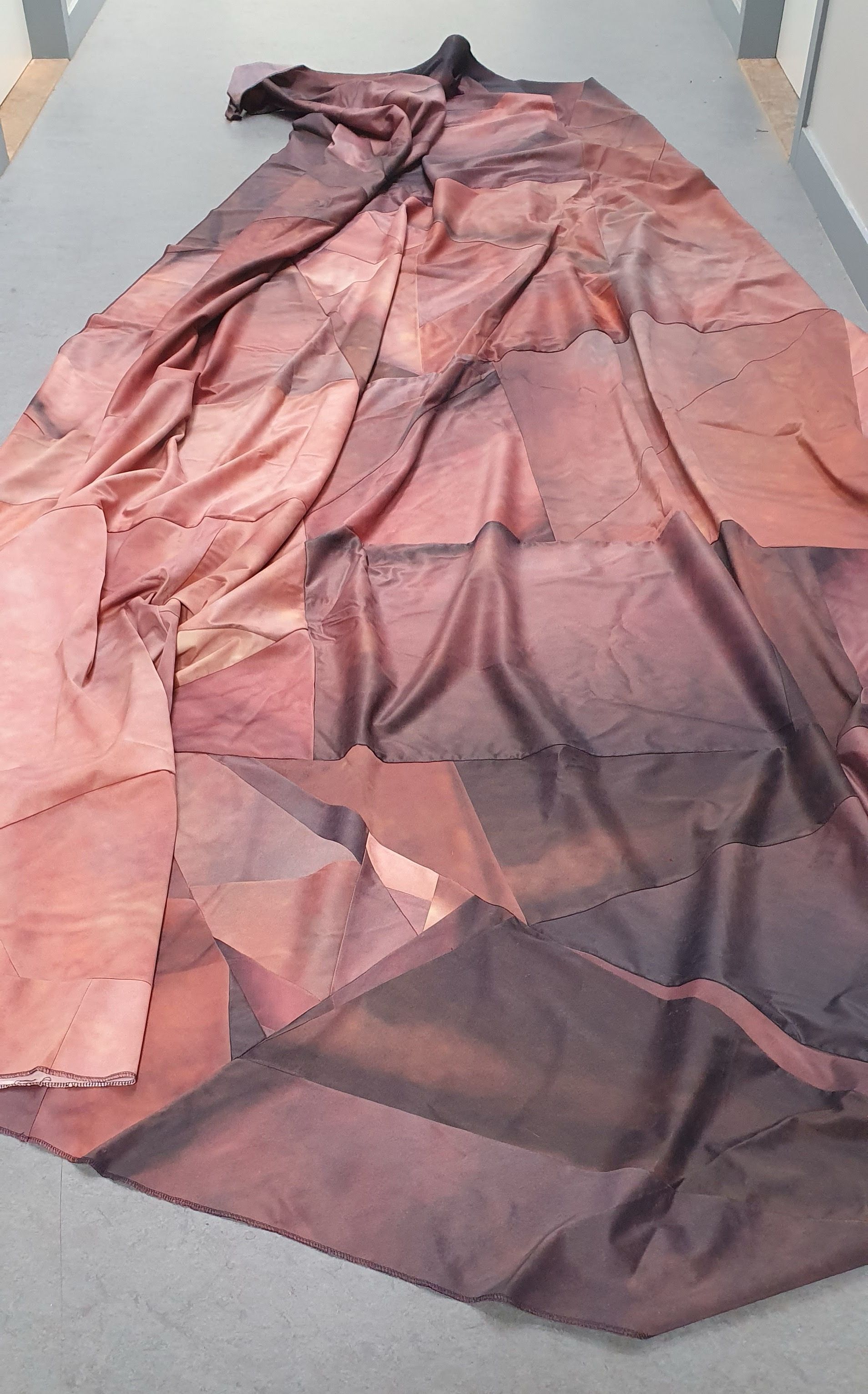 Op een grijs oppervlak ligt een lang, gevouwen textiel met een geometrisch patchwork in de kleuren bruin, oranje en rood.