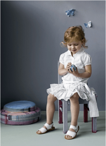 Meisje in een witte papieren outfit zit op een stoel en houdt een voorwerp vast. Twee papieren vlinders aan de muur.